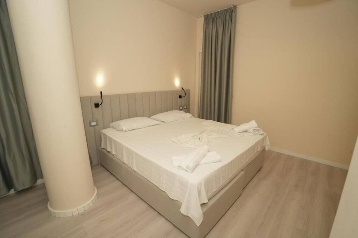 Un letto rifatto con lenzuola bianche e un asciugamano arrotolato, una testiera di colore chiaro e un lampadario fissato al muro.