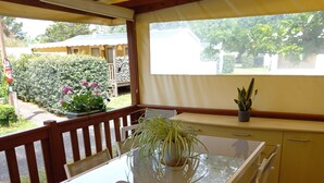 A covered patio with a glass-top table and white chairs, a small potted plant, and a view of a garden and house in the background.