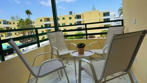Ein Balkon mit vier weißen Stühlen, einem kleinen runden Tisch und einem Blumentopf. Der Balkon bietet Blick auf ein Wohngebiet mit gelben Gebäuden und Palmen.