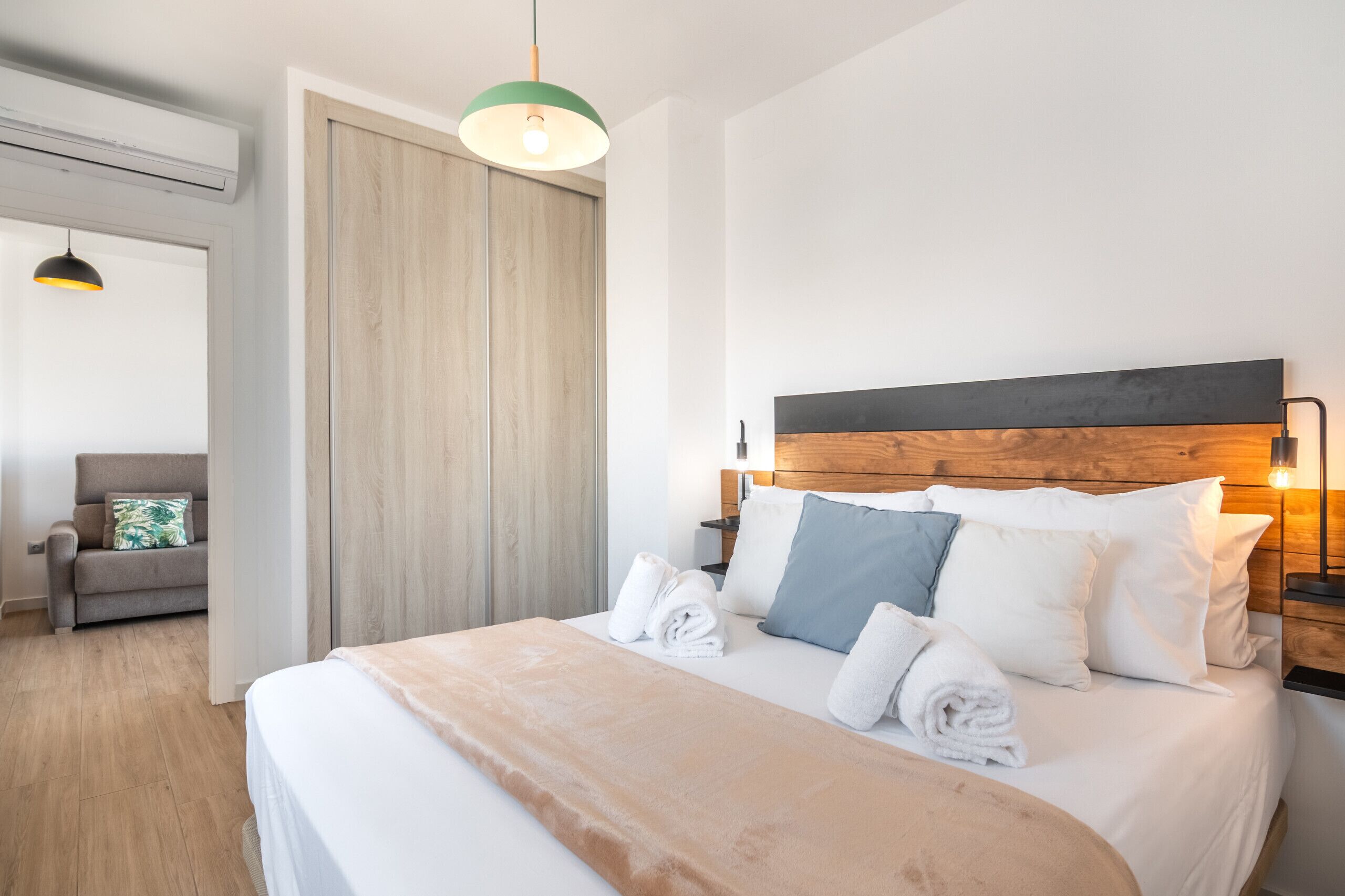 A modern hotel room with a wooden headboard, a large bed with white linens, a grey sofa, and a green pendant light.