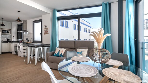 Una sala de estar moderna con una mesa de centro de vidrio, un sofá y un comedor con sillas. Se ve una cocina al fondo.