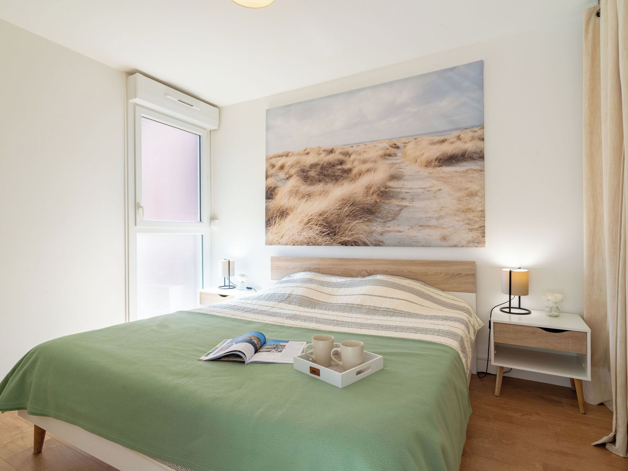 A bedroom with a bed, a nightstand, a lamp, a window, and a painting of a beach scene.