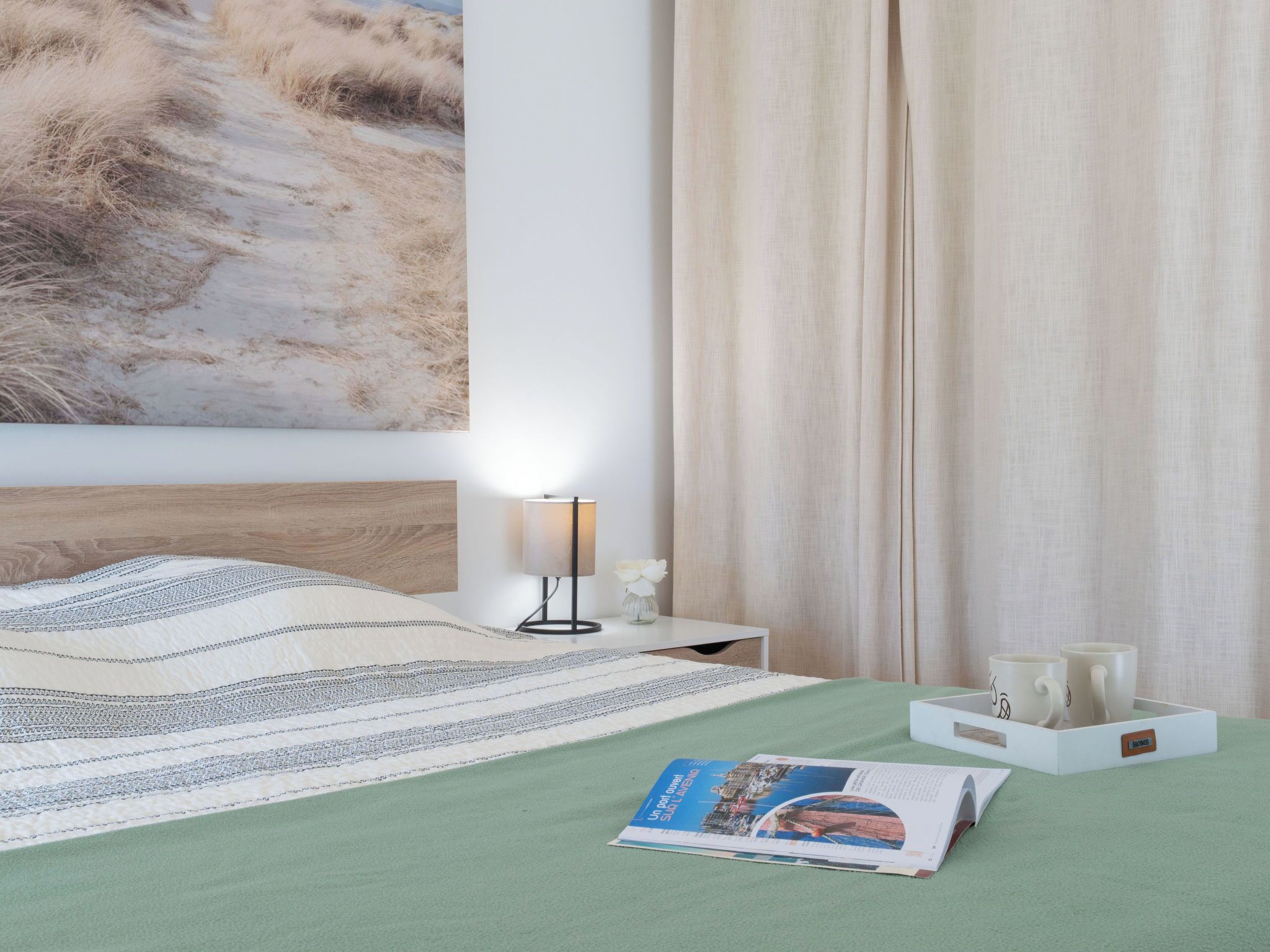 Una cama bien hecha con una colcha verde, una lámpara de noche, una revista y una bandeja con dos tazas.