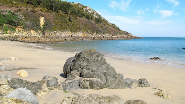 A beach with large rocks in the foreground, a sandy shore, and a rocky hillside with sparse vegetation. - Ti Breizh by Interhome (Guimaëc)