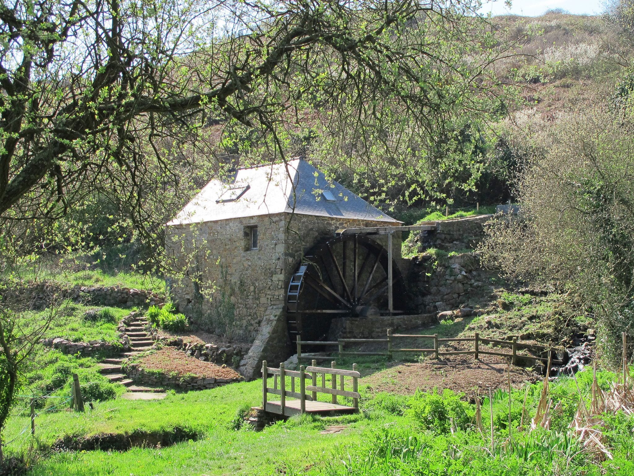 A stone watermill with a wooden wheel, surrounded by lush greenery and a stone staircase leading up to it.