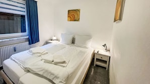 Habitación de hotel con cama doble, dos mesitas de noche, un radiador y una obra de arte colgada en la pared. Tiene además una ventana con cortinas.
