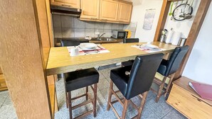 Cocina compacta con armarios de madera, microondas y un lavamanos. Cuatro sillas negras rodean una mesa de madera con un mantel individual.