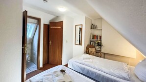 A small, cozy bedroom with two beds, a wooden wardrobe, a bathroom, and a small sitting area with a chair and a table. - Vacation home Tinnum by Interhome (Sylt)