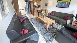 A modern living room with a black leather sofa, a wooden coffee table, and a green sofa with red cushions. A kitchen is visible in the background. - Vacation home Tinnum by Interhome (Sylt)