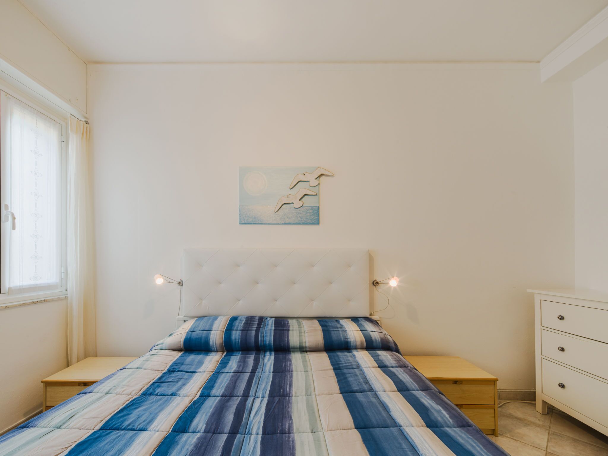 A bedroom with a bed, bedside tables, a nightstand, a dresser, a window with curtains, and a wall-mounted artwork.