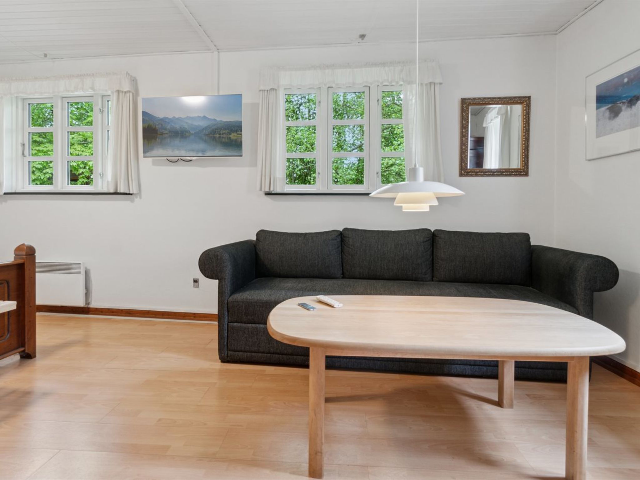 Ett rum med en mörkgrå soffa, ett träbord och två fönster med vita gardiner.