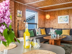 A room with a wooden ceiling, a sofa with colorful cushions, a table with a bottle and glasses, and a window with a view.