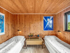 Ein Zimmer mit Holzwänden und -decke, zwei Einzelbetten, einem kleinen Tisch mit einer Lampe und einem Fenster.
