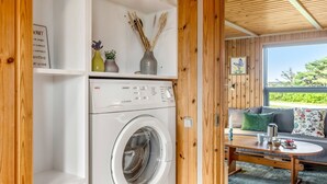 A laundry area with a front-loading washing machine, wooden shelves with decorative items, and a view of an outdoor seating area with a table and cushions.
