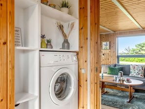 A laundry area with a front-loading washing machine, wooden shelves with decorative items, and a view of an outdoor seating area with a table and cushions.