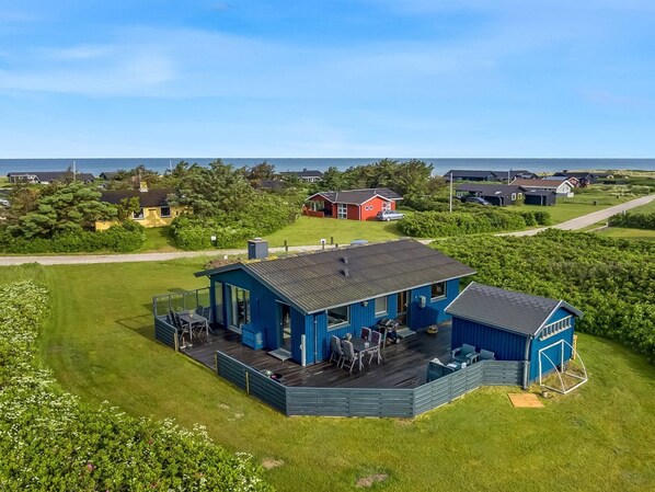 Ein blaues Gebäude mit einer Terrasse, umgeben von Grün und Blick auf das Meer.