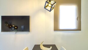 Un coin repas avec une table sombre, des chaises blanches, un centre de table composé d’un bol blanc, un luminaire fixé au mur et une œuvre d’art encadrée.