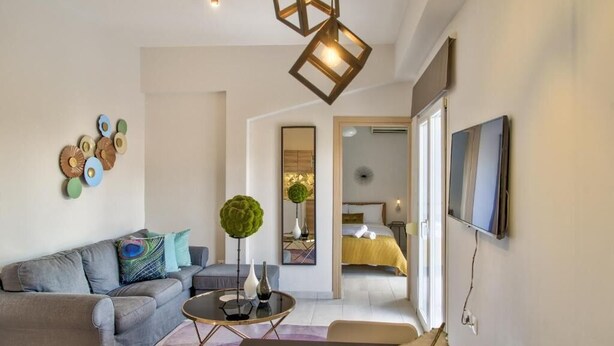 Un salon moderne avec un canapé, une table basse et des objets décoratifs. On aperçoit un lit au fond, avec vue sur l'extérieur.