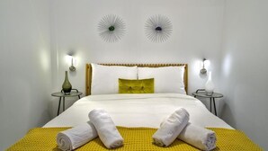 Un lit bien fait, avec du linge de lit blanc et un élément décoratif jaune, deux lampes murales décoratives et une petite table avec un vase de chaque côté.