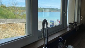 Une fenêtre de cuisine donnant sur une zone côtière, avec un mur en pierre et un robinet en métal sur le rebord de la fenêtre.