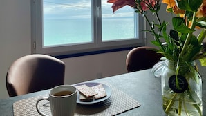 Une table avec une tasse, une assiette de nourriture et un vase de fleurs, près d’une fenêtre donnant sur la mer.