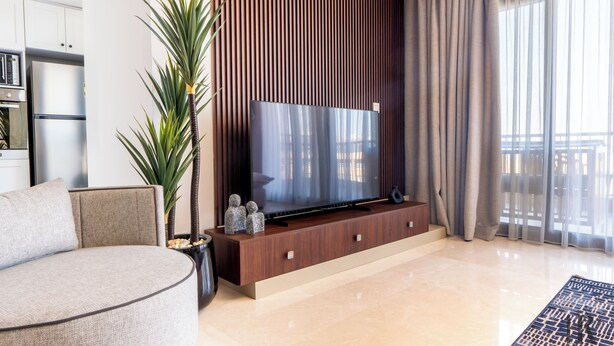 A modern living room with a flat-screen TV on a wooden stand, a sofa, a potted plant, and a large window with curtains. - Easy Rental/ Serviced Apartment In CFC New Cairo (NEW Cairo Governorate)