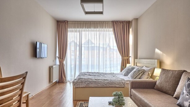 A modern hotel room with a large bed, a sofa, a TV, and a view of the city through sheer curtains. - MaRKoS Flat in Block 2, New Gudauri (Gudauri)