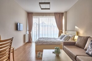A modern hotel room with a large bed, a sofa, a TV, and a view of the city through sheer curtains.