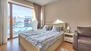 A bedroom with a large bed, a nightstand, a lamp, and a view of buildings and mountains through a sliding door. - MaRKoS Flat in Block 2, New Gudauri (Gudauri)