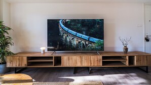 A modern living room with a large flat-screen TV mounted on a white wall, a wooden entertainment unit below, a potted plant to the left, and a vase with branches on the right. - City Center Modern Boho Stay (Nicosia)