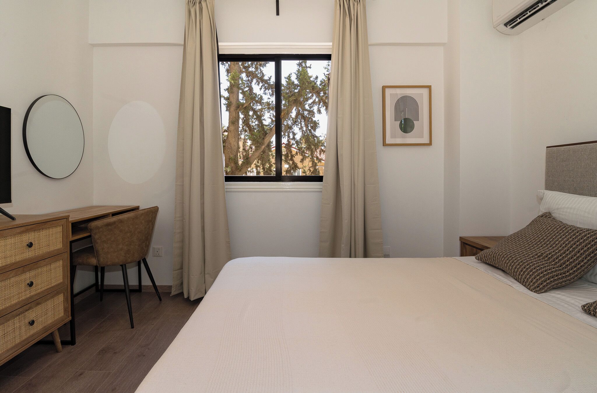 Un dormitorio con cama, escritorio, silla, cómoda, ventana con cortinas, y un cuadro en la pared.