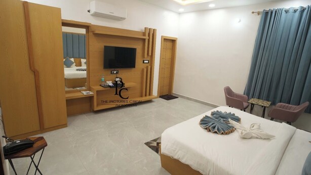 A hotel room with a bed, a TV mounted on a wooden panel, a desk with a phone, and a seating area with chairs. - Hotel Govind Residency (Mathura)