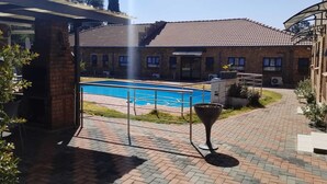 A swimming pool with a tiled deck, a brick building with a tiled roof, and a covered area with a metal roof. - Emperor Lodge And Tours (Germiston)