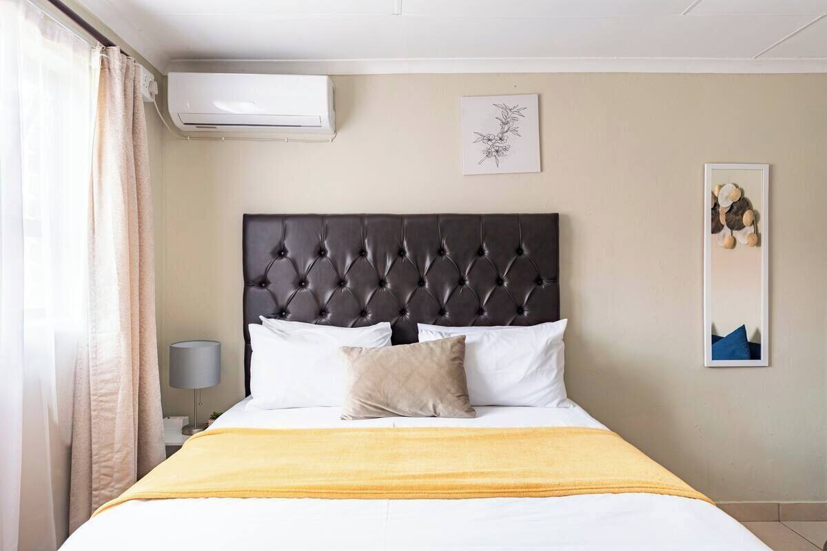 Ein Hotelzimmer mit einer gesteppten Kopfteil, einem Bett mit weißer und beiger Bettwäsche, einer gelben Decke und einer Wand-Klimaanlage, sowie zwei gerahmten Kunstwerken.