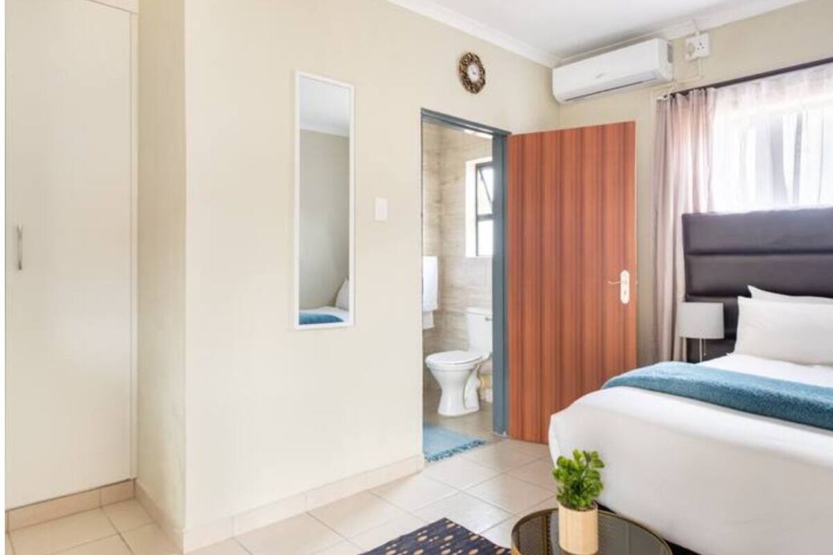 Ein Hotelzimmer mit einem Bett, einem kleinen Tisch mit Pflanze, einem Spiegel, einem Kleiderschrank und einem durchgegangene Tür erkennbaren Badezimmer.