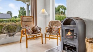 A cozy room with a fireplace, two wooden chairs with cushions, a floor lamp, and a view of greenery outside.