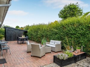 Une terrasse extérieure avec des meubles en osier, une table et des chaises. On aperçoit un abri de jardin noir à l’arrière-plan et une haie qui entoure l’espace.