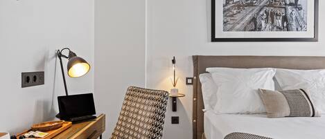 A hotel room with a bed, a desk with a laptop, a chair, a bedside table with cups and a tray, and a framed picture on the wall.