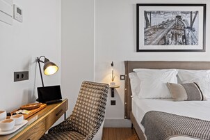 A hotel room with a bed, a desk with a laptop, a chair, a bedside table with cups and a tray, and a framed picture on the wall.