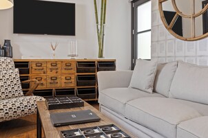 Een moderne woonkamer met een bank, een gestreept fauteuil, een houten salontafel met elektronische apparaten, een aan de muur bevestigde televisie en een groot raam met uitzicht.