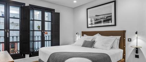 A modern bedroom with a large bed, a wooden headboard, a framed picture on the wall, and a window with a view of a building.