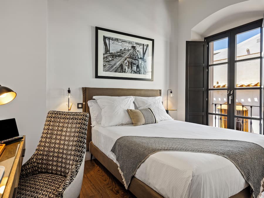 Una cama bien hecha con sábanas blancas y un plaid estampado, una silla con diseño geométrico, un escritorio con una computadora portátil y una taza, y una fotografía en blanco y negro enmarcada en la pared.