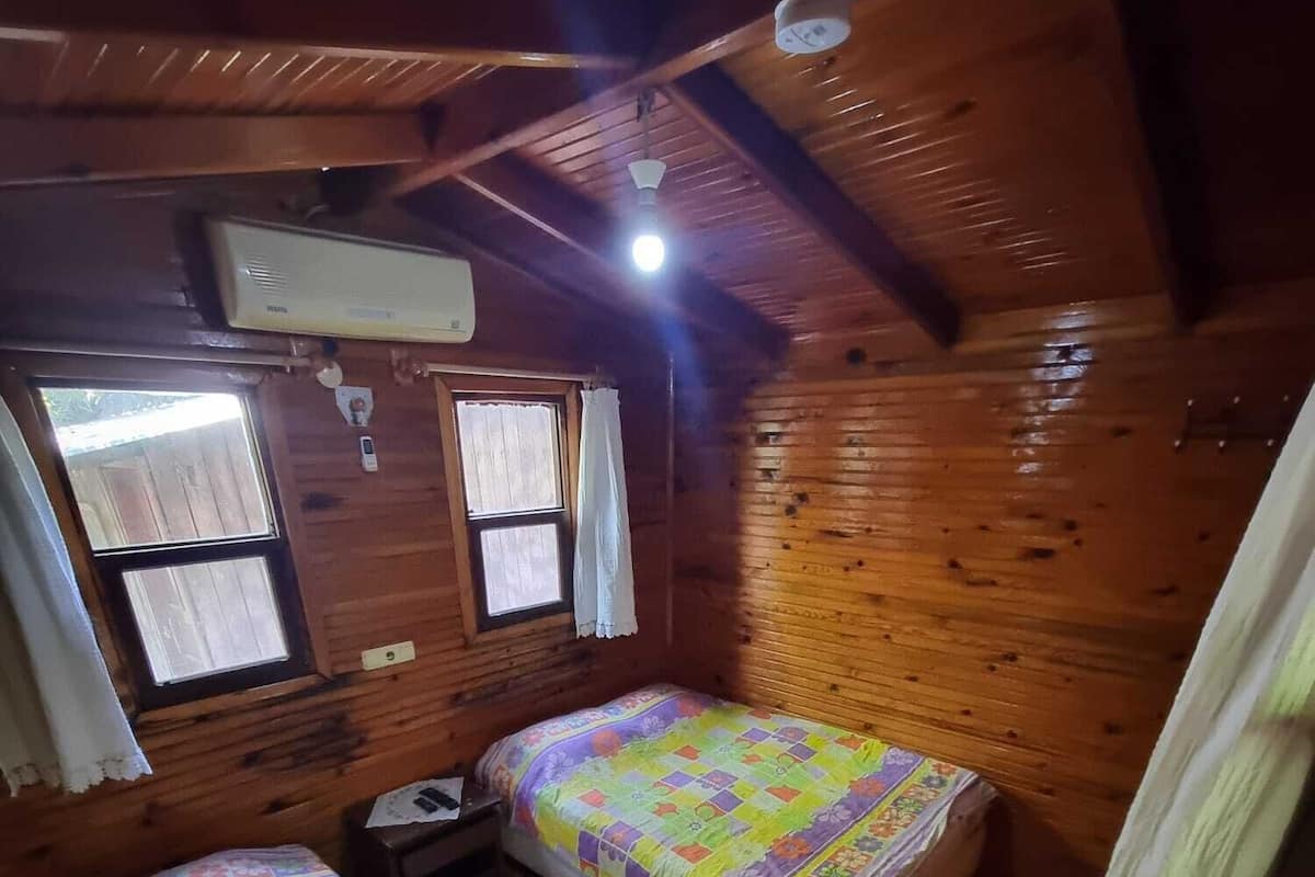 Uma cabana com duas camas, uma janela com cortinas, um ventilador de teto e uma mesa pequena.