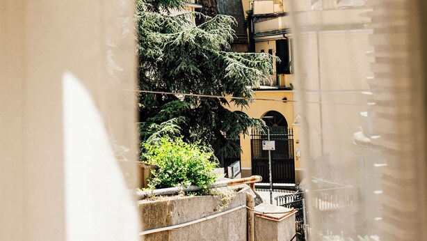 A view through a window with a curtain, featuring a stone bridge, greenery, and buildings with balconies. - Principe Genoa Home Train Station Piazza Principe (Unknown)