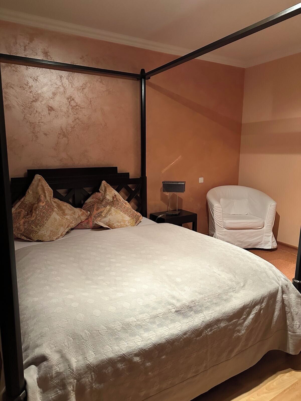 Un llit amb cortines, un edredó estampat, una tauleta de nit amb una làmpada i una butaca blanca en una habitació amb parets de color melocotó.