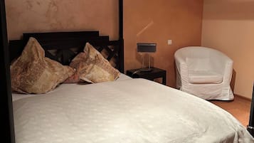 Un llit amb cortines, un edredó estampat, una tauleta de nit amb una làmpada i una butaca blanca en una habitació amb parets de color melocotó.