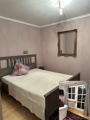 Un dormitorio con cama, mesita de noche, espejo, ventana con cortinas y un radiador.