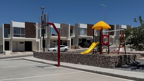 Ein modernes Gebäude mit einem Basketballplatz davor, einem Spielplatz mit einer gelben Rutsche und einer roten und gelben Spielkonstruktion.