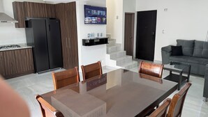 Eine moderne Küche mit Einbauherd, einem schwarzen Kühlschrank und Holzschränken. Es gibt einen Essbereich mit einem Esstisch mit Glasplatte und Holzstühlen. Ein Flachbildfernseher ist an der Wand über der Küche angebracht.