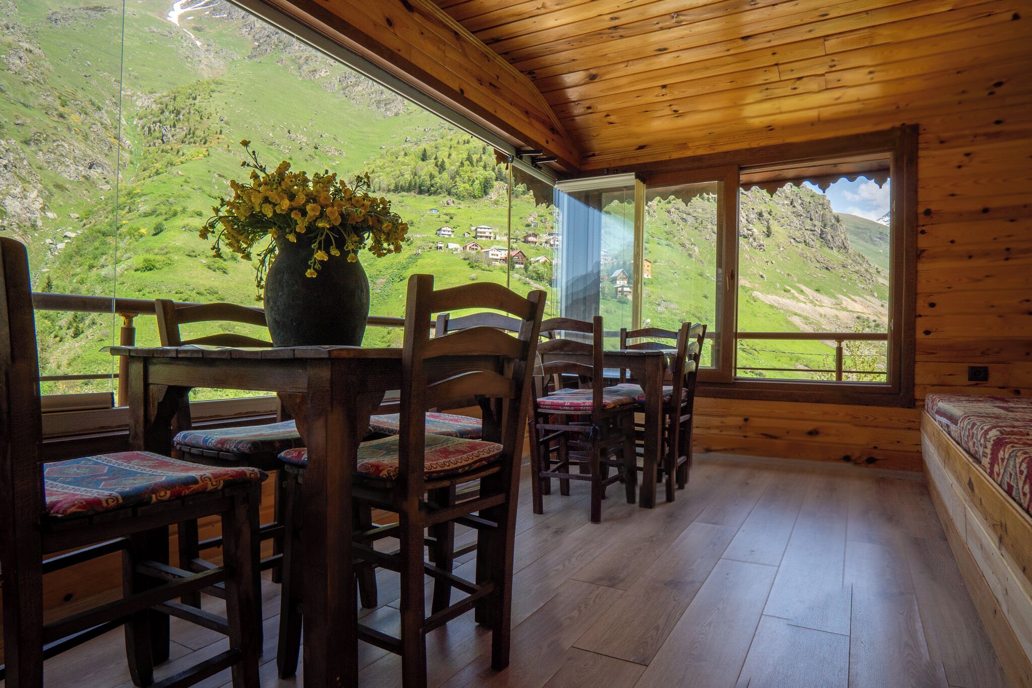 A wooden interior with a table and chairs, a vase with flowers, and a view of a green valley through large windows.